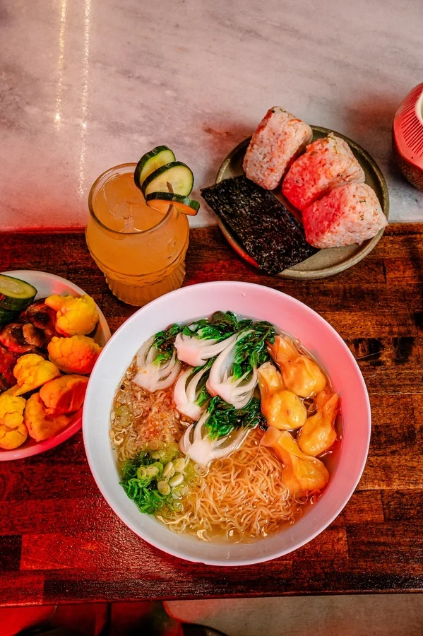 A bowl of Bareo wonton noodle soup with bok choy and a cocktail