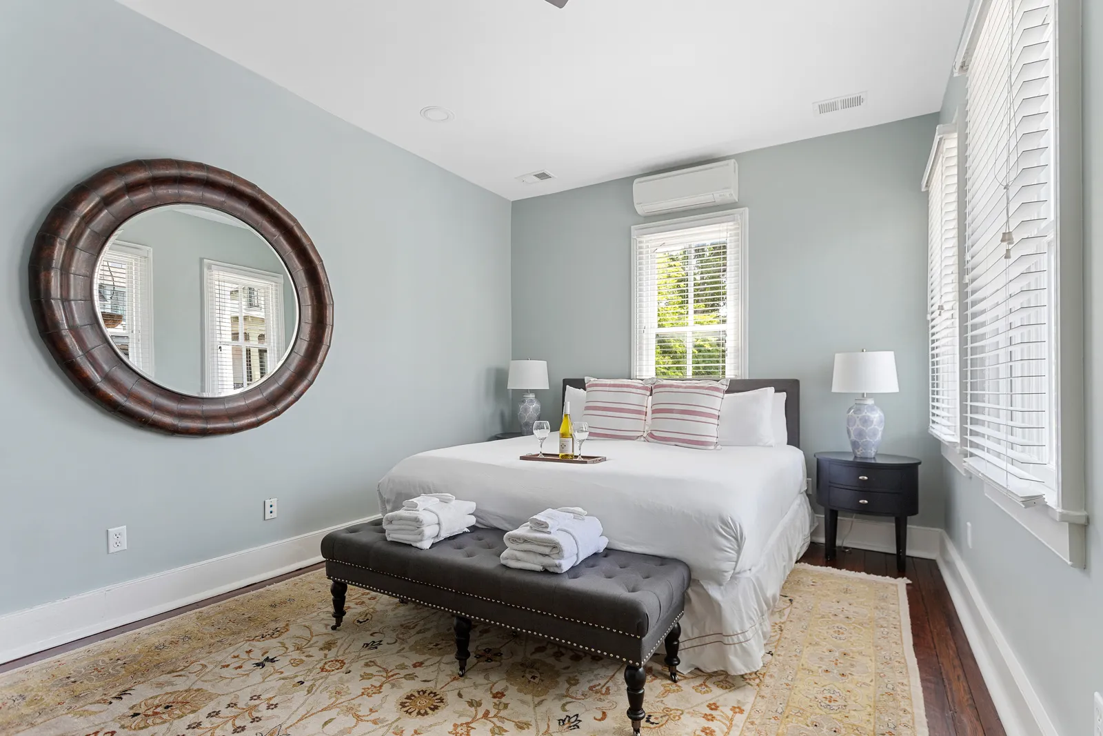 A queen bedroom with a round mirror and tufted bench